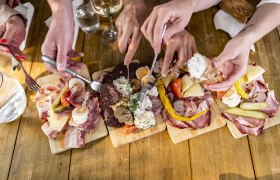 Mehrere H&auml;nde greifen nach Wurst- und K&auml;seplatten auf einem Holztisch.