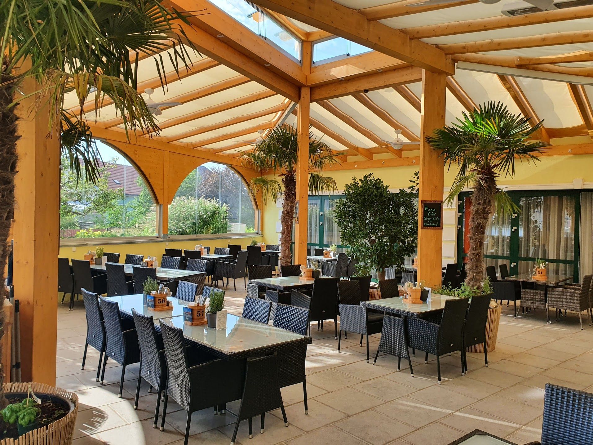 Überdachte Terrasse eines Restaurants mit Palmen und Tischen.
