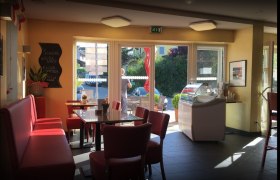 Innenbereich einer Bäckerei mit roten Sitzbänken und Tischen, großen Fenstern und einer Eistheke.